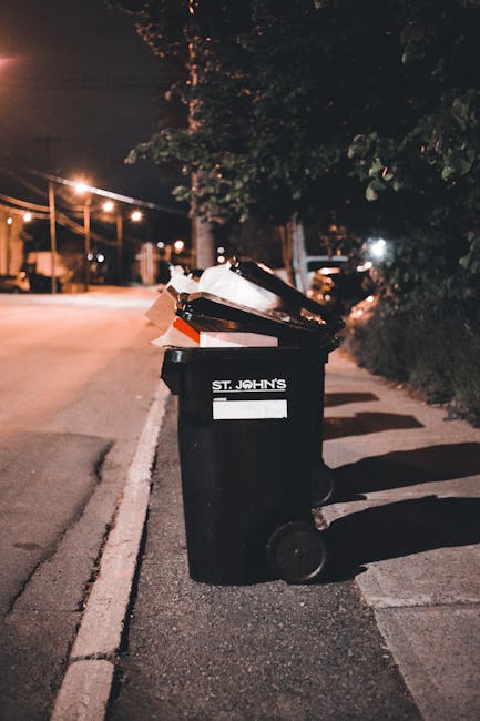 A black wheelie bin labeled 'St. John's' placed on a pavement next to a curb during nighttime. The bin contains discarded cardboard boxes, paper, and plastic packaging materials, some of which are protruding from the open lid. The scene is illuminated by streetlights, with a quiet residential street visible in the background, featuring trees, a parked car, and houses. The image captures the initial stage of waste disposal related to house moving or packing activities, as part of scheduled rubbish collection prior to a home relocation, exemplified by the services provided by Man with Van Sydenham. The scene emphasizes the importance of proper waste management in the context of household removals and furniture transport, with a focus on logistics and packing procedures involved in an efficient move.