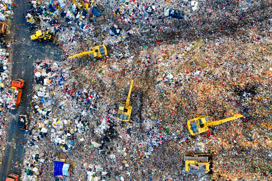 An aerial view of a large landfill site filled with a vast accumulation of mixed waste, including plastic bags, cardboard boxes, textiles, and other debris. Several yellow excavators with long arms are actively engaged in moving and digging through the waste, with one near the center lifting materials and others positioned around the site. A section of a paved road runs along the left side of the image, with smaller vehicles and waste containers situated nearby. The surrounding area is covered in waste, and the scene depicts the scale of waste management operations, relevant to home relocations and disposal services offered by Man with Van Sydenham as part of their house removals and waste disposal solutions.