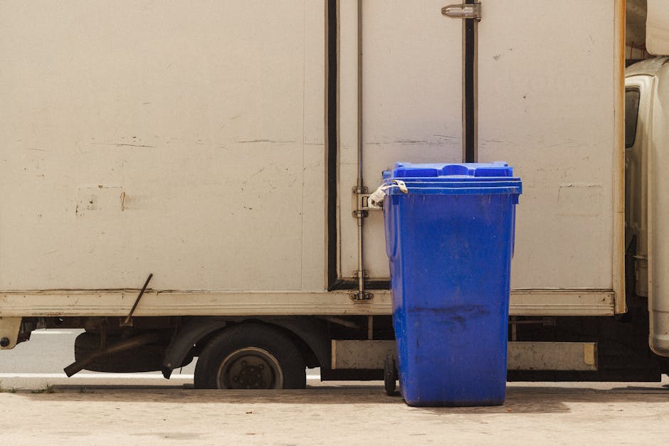A black wheelie bin labeled 'St. John's' placed on a pavement next to a curb during nighttime. The bin contains discarded cardboard boxes, paper, and plastic packaging materials, some of which are protruding from the open lid. The scene is illuminated by streetlights, with a quiet residential street visible in the background, featuring trees, a parked car, and houses. The image captures the initial stage of waste disposal related to house moving or packing activities, as part of scheduled rubbish collection prior to a home relocation, exemplified by the services provided by Man with Van Sydenham. The scene emphasizes the importance of proper waste management in the context of household removals and furniture transport, with a focus on logistics and packing procedures involved in an efficient move.