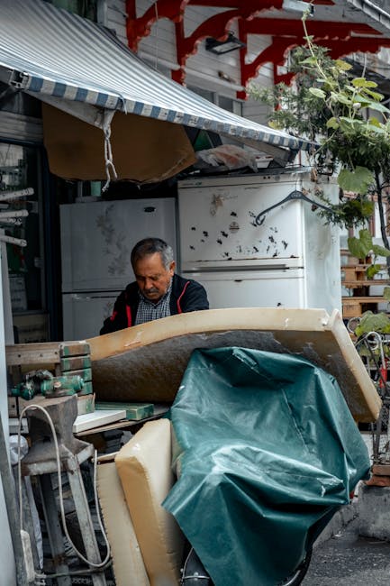 A man with a professional uniform is working outdoors near a residential building, preparing to load or unload large household items for a home relocation. In front of him, there is an old sofa partially covered with a green protective sheet, positioned on a wheeled trolley or moving equipment, with some packing materials visible around. Behind him, a white fridge and other household furniture and boxes are arranged in a small driveway or loading area, with a blue-and-white striped awning overhead providing partial shelter. The scene includes visible plants and foliage, suggesting an outdoor setting, and the environment is well-lit, likely during daytime. This image illustrates furniture transport and packing operations typical of professional removals, highlighting the careful handling of bulky items associated with house removal services provided by Man with Van Sydenham.