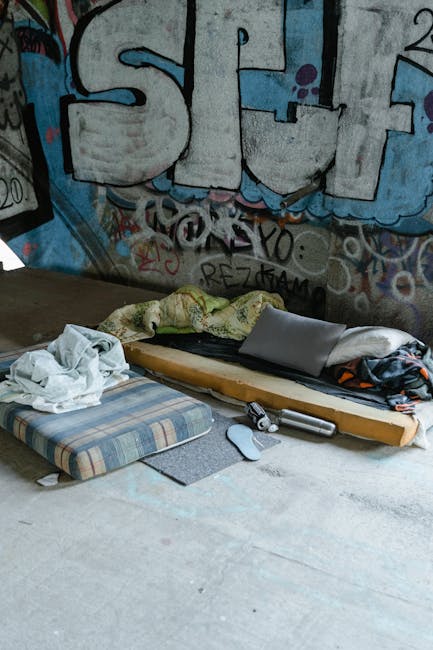 A bright blue chaise lounge with two matching cushions is positioned on the sidewalk outside a row of shopfronts in an urban environment. The furniture is made of fabric upholstery with wooden legs and appears to be ready for removal or transport. Behind it, there are multiple closed storefronts with metal roller shutters, some covered in graffiti, including signs for a clothes shop, a furniture store, and a pharmacy. The shopfronts are part of a brick building with multiple windows on the upper floors. The scene is during daylight hours, with an overcast sky visible at the top of the image. The setting suggests a home relocation or furniture transport process, with the furniture placed on the pavement outside for collection by a professional moving service such as Man with Van Sydenham, operating within the removals sector.