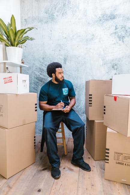 A man with dark, curly hair and a beard, dressed in a red long-sleeve shirt, stands in front of a plain light grey wall. He is holding a cardboard box with a large white label that reads 'OFFICE' on t