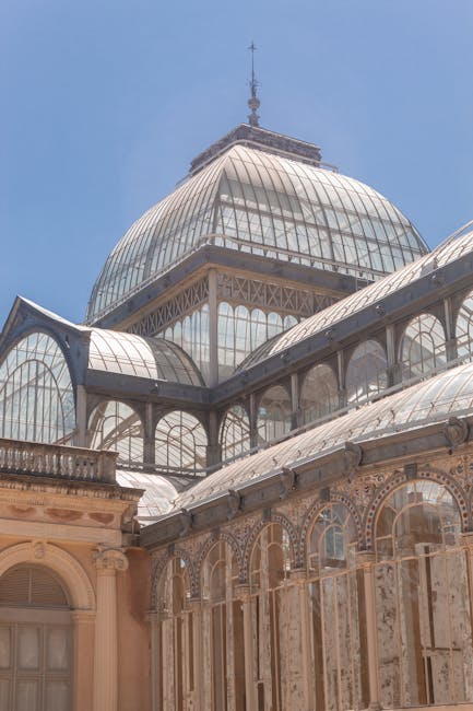 A detailed exterior view of a large historic glass and metal conservatory with ornate stonework seen from ground level, capturing the intricate iron framework supporting curved glass panels that form a prominent domed structure beneath a tall spire on top. Part of the conservatory's lower section features classical architectural elements including arched windows and decorative stone carvings. The lighting highlights the transparency of the glass, and the clear blue sky serves as a backdrop. This image relates to home relocation and packing processes managed by Man with Van Sydenham, illustrating the scale and complexity of transporting and handling large, fragile glass structures during a house move or furniture transport, with an emphasis on careful loading and logistical planning.