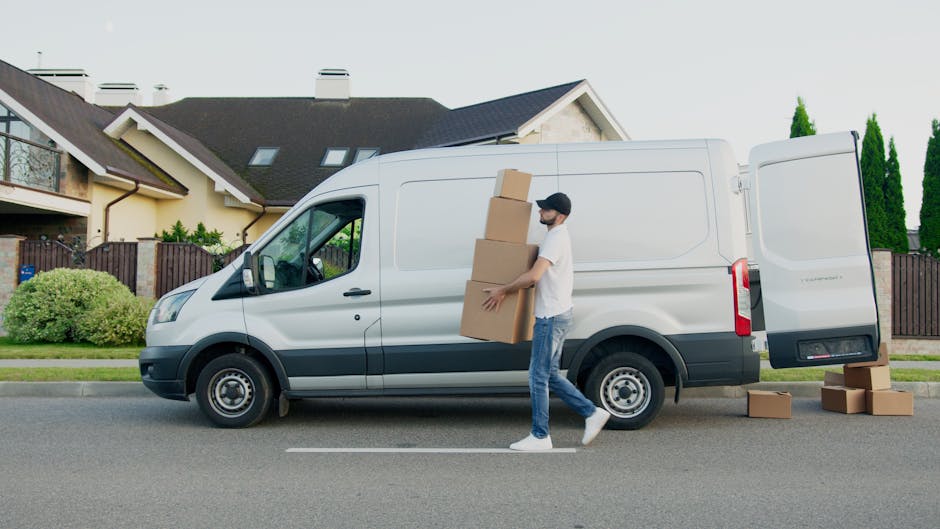 A man wearing a white T-shirt, jeans, white sneakers, and a black cap is outdoors on a residential street, loading three cardboard boxes into the side of a white panel van with its sliding door open. 