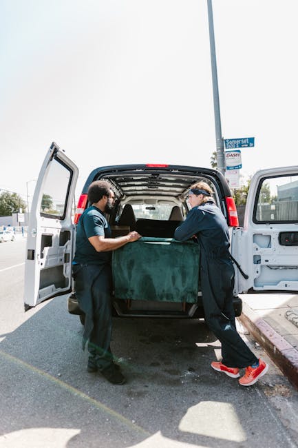 A man wearing a white T-shirt, jeans, white sneakers, and a black cap is outdoors on a residential street, loading three cardboard boxes into the side of a white panel van with its sliding door open. 
