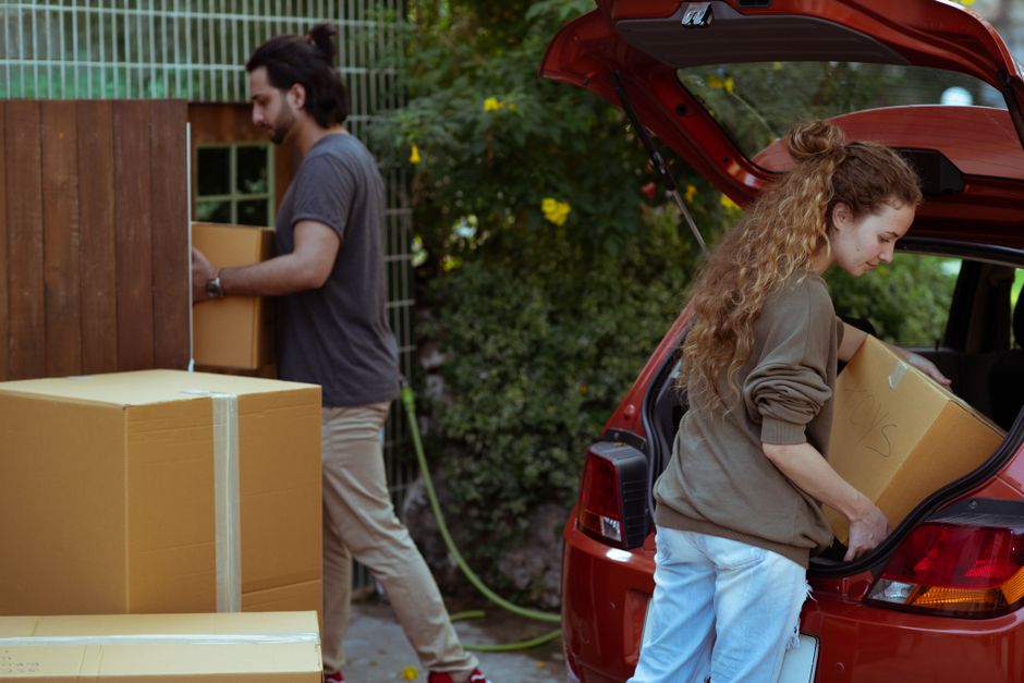 A man with dark hair tied back, wearing a grey t-shirt and beige trousers, is seen lifting and carrying a small cardboard box towards a wooden gate with vertical slats, set outdoors in a garden environment. Behind him, there are larger cardboard boxes stacked on the ground, wrapped with packing tape. To the right, a young woman with long, curly red hair, dressed in a brown jacket and light blue jeans, is placing a medium-sized cardboard box into the open trunk of a red hatchback vehicle parked on a paved driveway. The interior of the car's trunk is visible, showing additional packed boxes. In the background, leafy green bushes and trees add natural surroundings, with sunlight illuminating the scene, indicative of a home relocation or moving process. The setting demonstrates the loading phase of a house removal, with packing materials, boxes, and equipment such as the car trunk involved in furniture transport and moving logistics, consistent with services provided by Man with Van Sydenham.