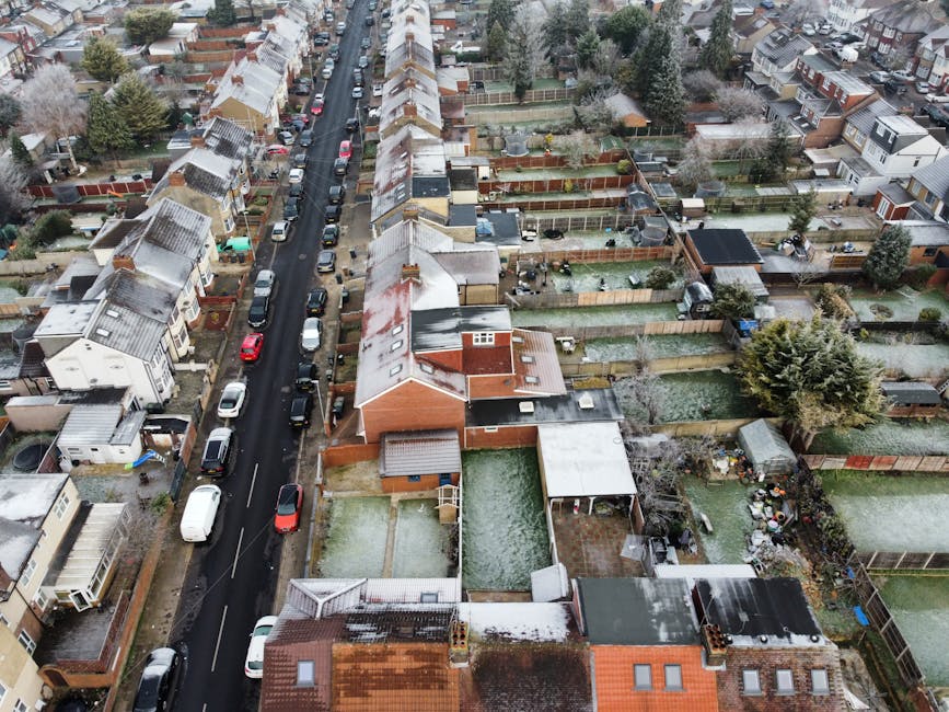 An aerial view of a residential street in Sydenham showing rows of terraced houses with pitched roofs, some with snow-covered surfaces. Parked cars line both sides of the narrow asphalt road, and the houses feature small front gardens enclosed by fences. The image reveals back gardens of varying sizes, some with garden furniture, sheds, or small outbuildings, and a few gardens are covered with light snow. Visible at the bottom center is a house with an adjacent backyard containing a patio area and a small garden. The overall scene depicts a typical suburban neighborhood during winter, with signs of home relocation preparations such as packed boxes and furniture being moved, consistent with professional house removal services by Man with Van Sydenham in the context of complying with Lewisham Council's moving rules regarding permits and kerbside loading.