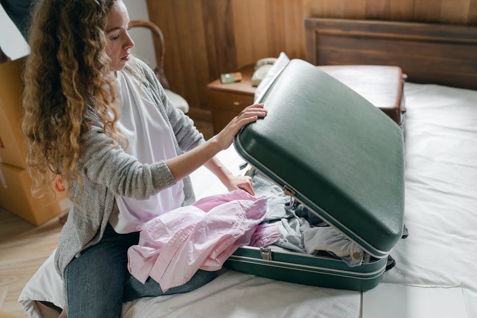 A woman with long, curly blonde hair, dressed in a light gray cardigan and white top, is sitting on a bed in a bedroom and preparing to pack a dark green hard-shell suitcase. She is holding the suitcase open with her left hand while placing pink and gray clothing inside with her right hand. The suitcase is partly filled with folded clothes and positioned on the bed, which has a white bedsheet. Behind her, there is a wooden headboard, a bedside table with a white telephone, a small object, and a wooden drawer. The room features wood-paneled walls, and natural light illuminates the scene, indicating daytime. This image relates to packing during a house move or home relocation, and the scene depicts the process of preparing personal belongings for transport as part of a professional removals service, such as that provided by Man with Van Sydenham.