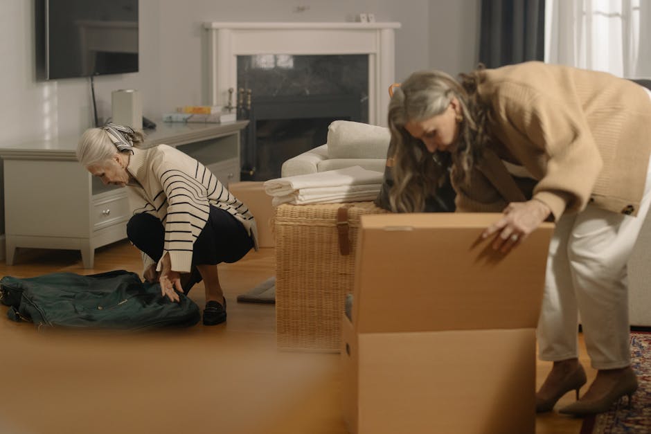Two women are in a living room engaged in packing and preparing for a home relocation. The woman on the left, with gray hair and wearing a striped long-sleeve top, is kneeling on the wooden floor, folding or organizing a green backpack or duffel bag. The woman on the right, with long gray hair and dressed in a beige coat and white pants, is bending over and lifting the lid of a cardboard box, likely packing or preparing items for transport. The room features a white chest of drawers, a white fireplace with a glass cover, and a modern TV mounted on the wall. There are neatly folded white towels or linens stacked on a wicker basket near the fireplace. The window in the background allows natural light to illuminate the space, underscoring the careful handling of possessions for a move. This scene depicts the packing process involved in furniture transport and home relocation, aligning with the services offered by Man with Van Sydenham as detailed in the packing and access guide for Thorpewood Avenue moves.