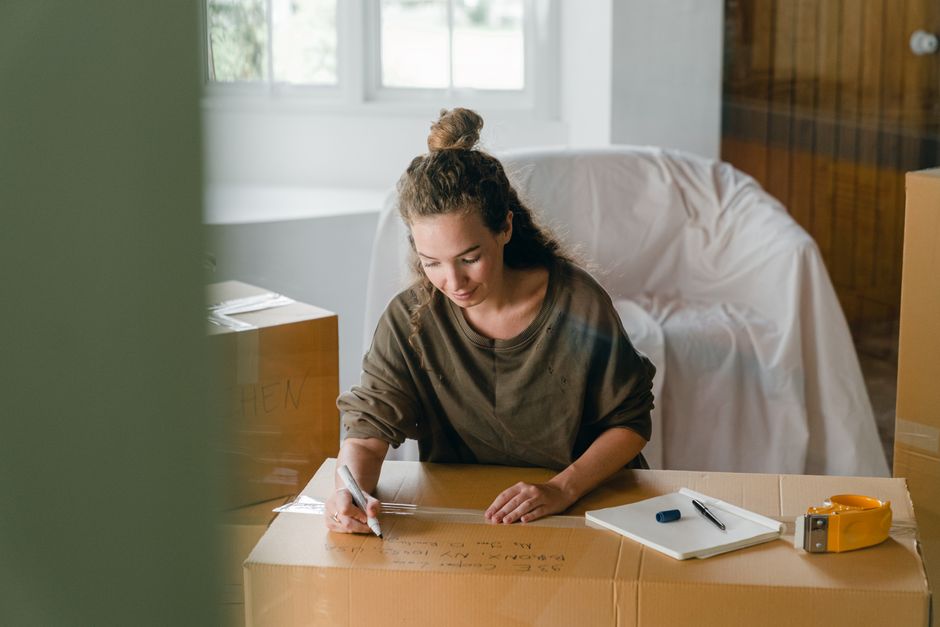A young woman with curly hair tied back in a bun is seen inside a residential property, sitting at a wooden table surrounded by packing materials. She is wearing a brown long-sleeved top and is focused on writing notes or instructions on a large cardboard box with a black marker. In front of her on the table are a white notepad, a black pen, and a yellow tape measure. Behind her, there are stacked cardboard boxes, some partially wrapped in plastic or fabric for protection during home relocation. The room has a window letting in natural daylight, illuminating the scene clearly. She appears to be involved in the packing and moving process, which may include labeling or organizing belongings for transportation. The image captures the preparation stage of furniture transport or house removals, with the company's services, Man with Van Sydenham, supporting professional packing and load planning, especially relevant for large-item moves in SE26, involving careful handling and logistical coordination.