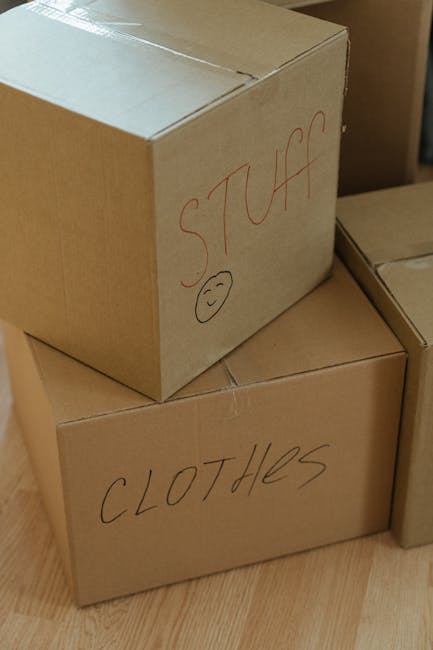 Close-up view of several cardboard boxes prepared for home relocation, placed on a wooden floor inside a property. The boxes are labeled with handwritten text: one box has 'STUFF' written in red marker, accompanied by a smiley face, while another box is labeled 'CLOTHES' in black marker. The boxes vary in size, with some stacked and others positioned nearby. The environment suggests an indoor setting during packing or loading for a move, with items secured with packing materials such as cardboard and markers used for labelling. The scene is lit with natural or ambient indoor lighting, capturing the typical process of furniture transport and packing for house removals, as handled by [COMPANY_NAME].