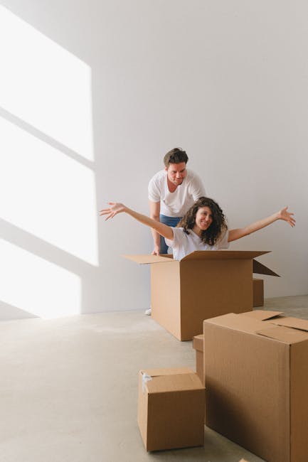 A young woman with curly hair is sitting inside a cardboard moving box with her arms raised and a joyful expression, while a man stands behind her, smiling and supporting her from behind; they are in 