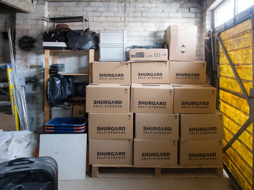 Inside a storage unit with a rough brick wall, a shelving unit holds black plastic bags and miscellaneous items, while a pallet stacked with nine uniformly-sized cardboard boxes labeled 'SHURGARD SELF-STORAGE' dominates the scene, indicating recent or ongoing home relocation or furniture transport activities. To the left, a metal ladder leans against the wall, and on the floor are folded collapsible trays and a piece of white foam board. The right side of the image features a partially visible yellow-tinted sliding door, suggesting an access point for loading or unloading household goods. The lighting is natural, coming through the partially open door, illuminating the packed items. This setting reflects typical storage solutions used during short-term moving or packing and moving processes facilitated by companies like Man with Van Sydenham, supporting logistics for house removals and furniture transport near SE26, Sydenham.