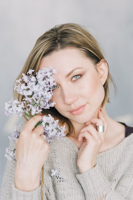 There is no relevant visual content related to house removals, moving services, or storage. The image shows a woman with shoulder-length light brown hair holding a small bouquet of light purple flowers close to her face, partially covering one eye. She is wearing a beige knitted sweater and has a neutral expression, with soft lighting highlighting her features against a plain, light-colored background. This image does not depict furniture, boxes, appliances, or any elements associated with relocation or packing processes, and does not involve any moving equipment or vehicles.